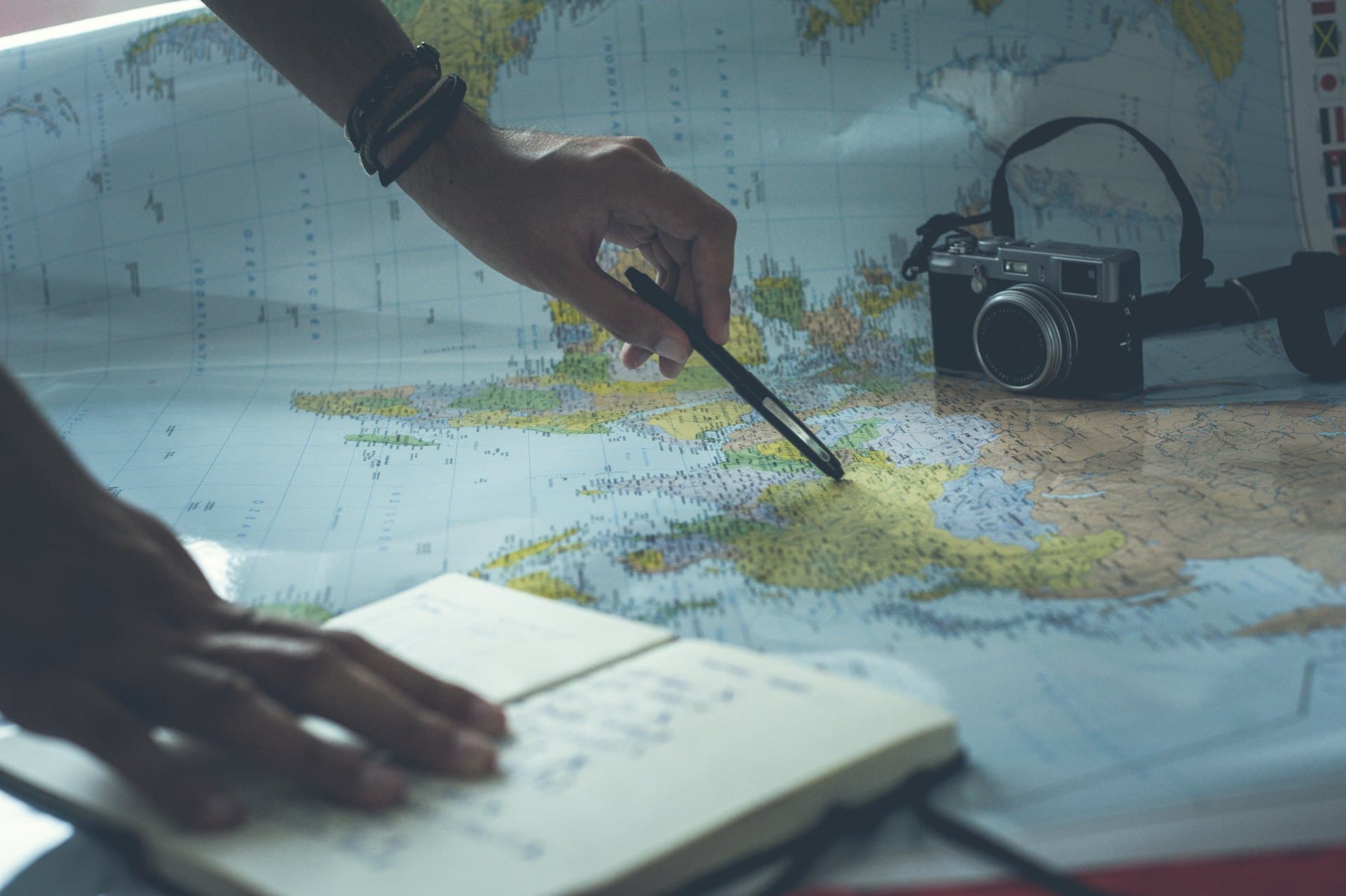 Une main trace un parcours sur une carte du monde, un carnet et un appareil photo à côté